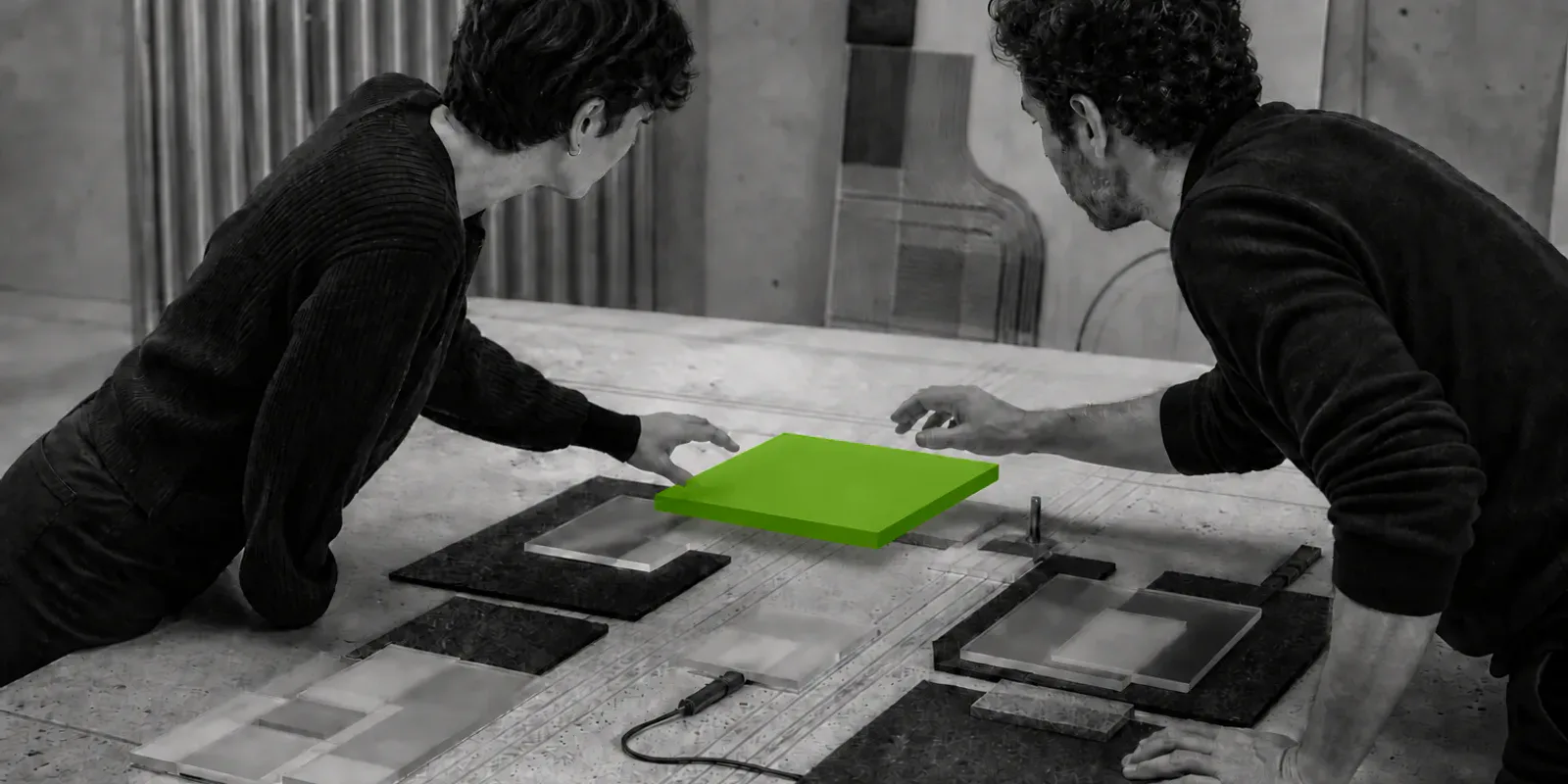 Dos personas en blanco y negro trabajan sobre una mesa compartida mientras un módulo verde flota entre sus manos.