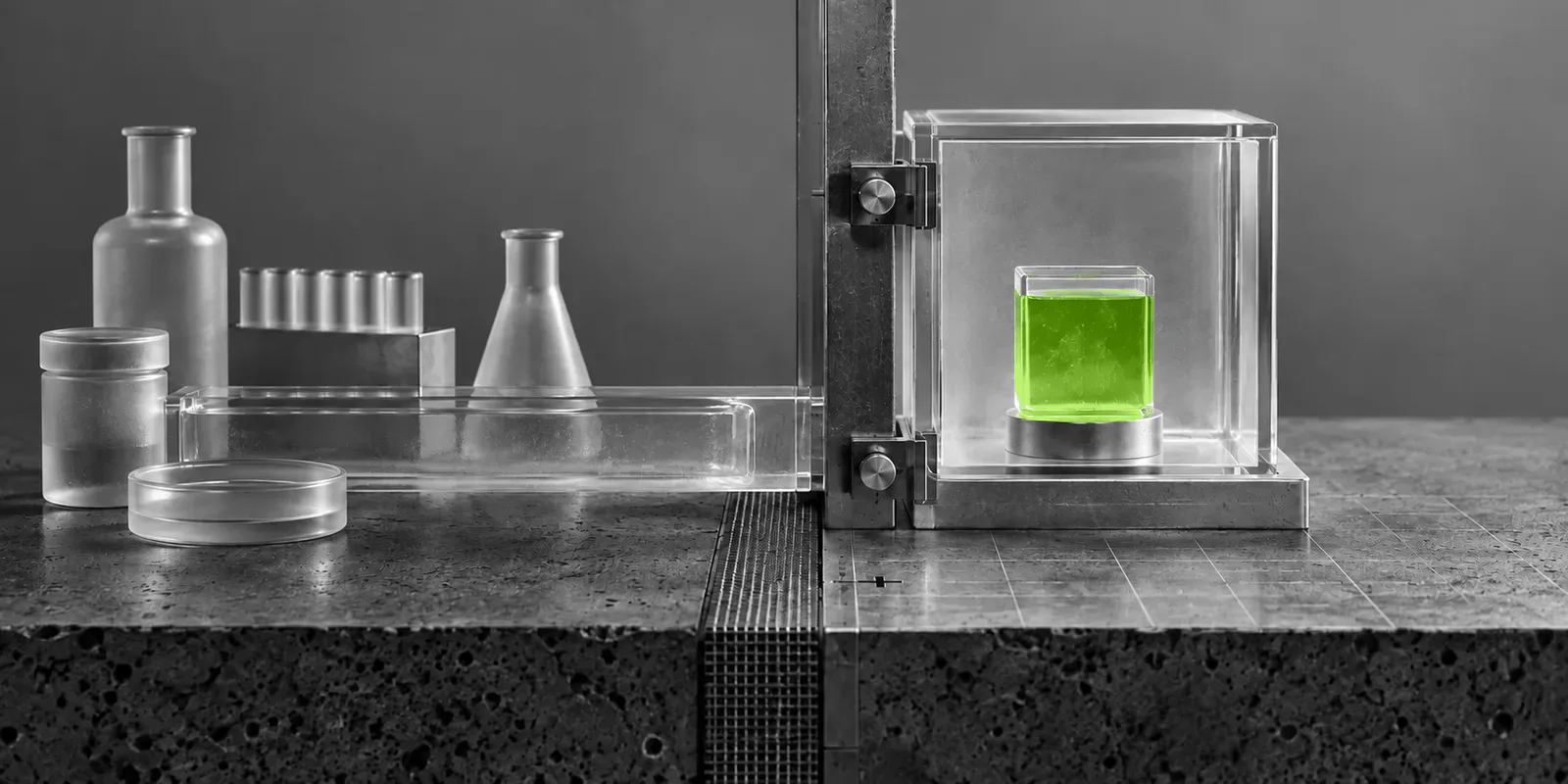 Una frontera metálica separa recipientes de laboratorio grises de un resultado verde contenido en una cámara transparente.