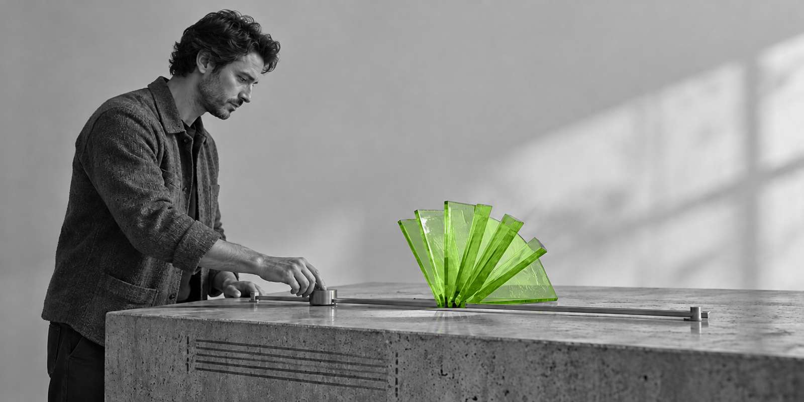 Una persona ajusta una pieza sobre una mesa de experimentación y modifica una pequeña estructura de vidrio verde, en una escena de hormigón y metal en blanco y negro.