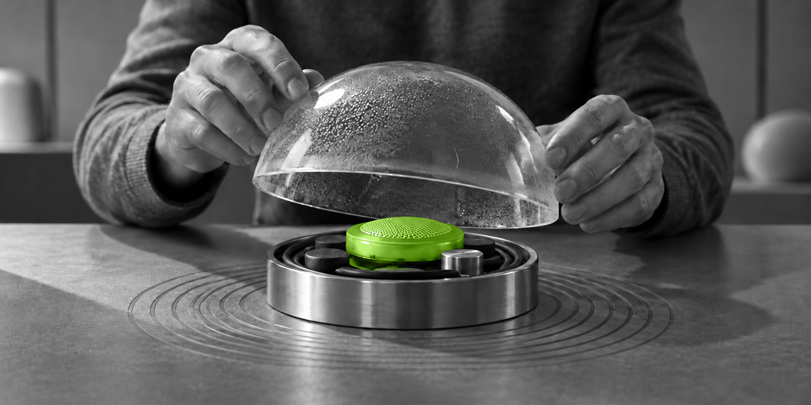 Fotografía conceptual en blanco y negro de una persona adulta anónima, en primer plano y con las manos visibles, levantando con cuidado una cúpula de vidrio empañado para revelar un pequeño dispositivo verde brillante sobre una base metálica y de goma, con anillos concéntricos grabados alrededor.