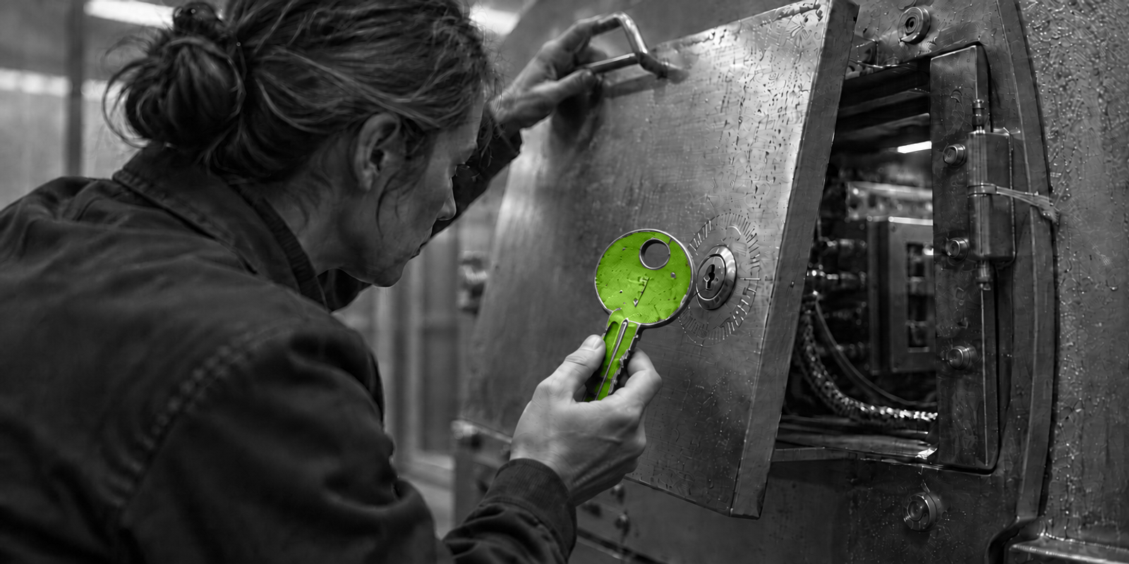 Fotografía documental en blanco y negro de una persona adulta anónima, en perfil izquierdo y parcialmente de espaldas, abriendo con una mano una tapa metálica de inspección mientras con la otra extrae una pequeña llave verde brillante de la cerradura; la escena muestra metal rayado, condensación y un motivo circular grabado alrededor del bombín.