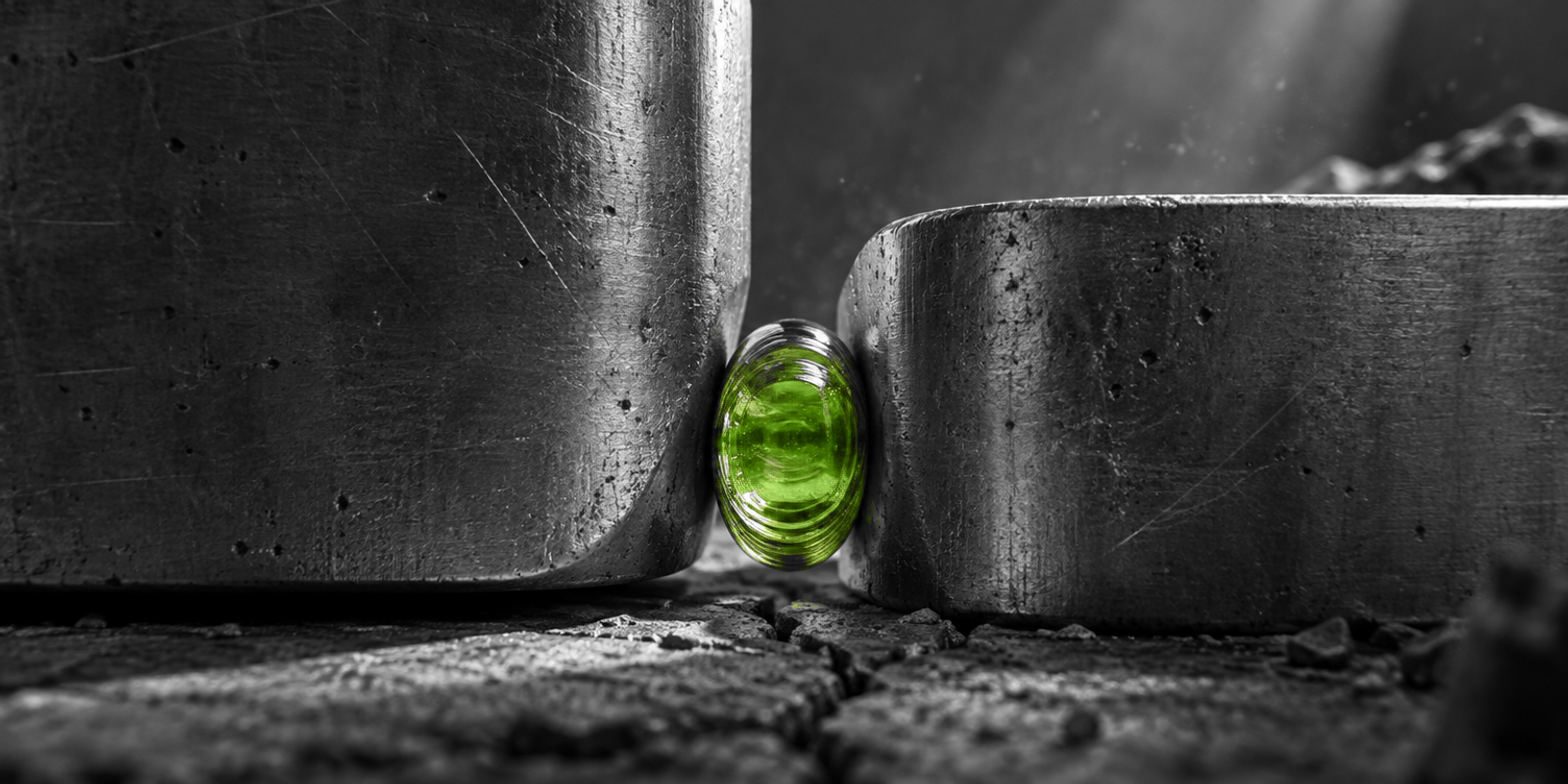 Fotografía conceptual en blanco y negro de dos masas industriales de metal cepillado y piedra agrietada, con una única pieza de vidrio verde translúcido, pequeña y comprimida en el centro. El objeto verde muestra estrías internas y surcos concéntricos, y actúa como punto focal entre ambas formas.
